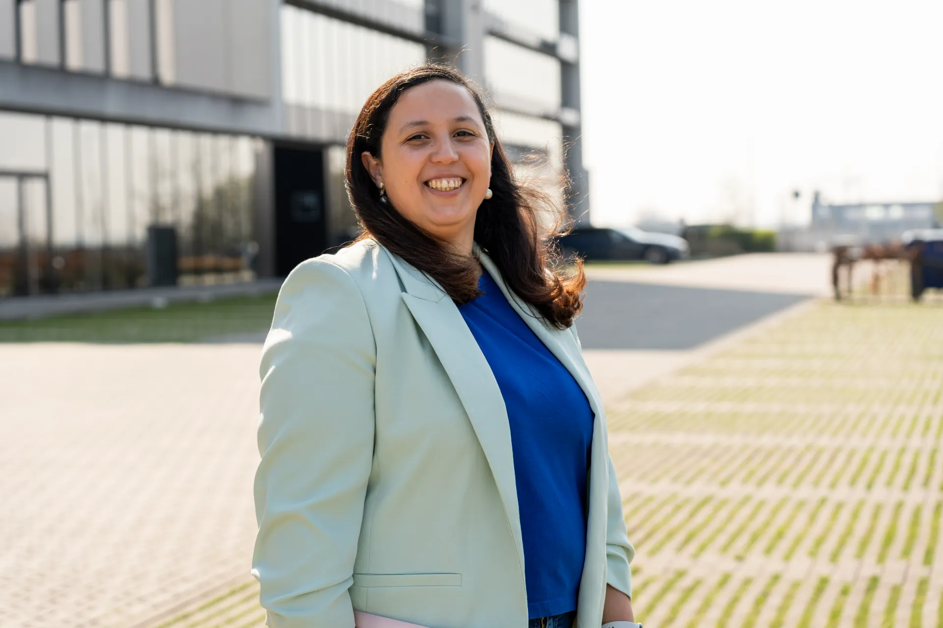 Financieel Advies KMO – Samra Daoud Lachende financieel adviseur Samra Daoud met lichte blazer buiten voor modern kantoorgebouw.